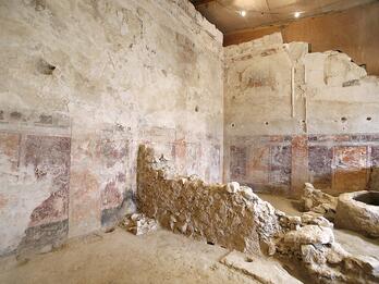 Ancient, weathered room with faded wall paintings, exposed stone walls, and a rough stone structure in the center.