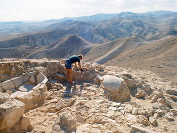 A person examines ancient stone ruins atop a rocky hill, surrounded by a vast, arid mountain landscape under a partly cloudy sky. Archaeological tools are scattered nearby.