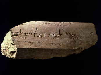 A weathered stone slab with an ancient Hebrew inscription carved into its surface, displayed against a black background. The stone is uneven and partially broken on the left side.
