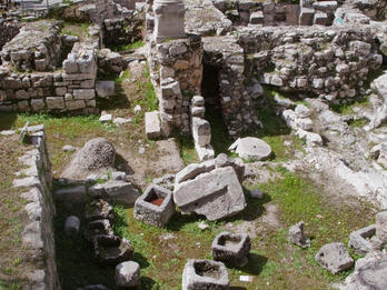 Ancient stone ruins with broken columns and scattered stone blocks, surrounded by moss and grass, set within ancient stone walls and partially preserved structures.