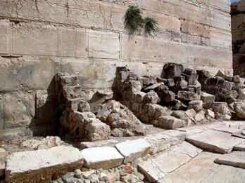 Ancient large stone blocks and rubble rest against a weathered stone wall with some sparse greenery.