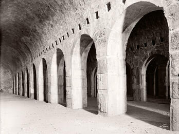 A black-and-white photo of a stone building interior shows a row of large, evenly spaced arched doorways under a vaulted ceiling, with light casting shadows on the sandy floor.