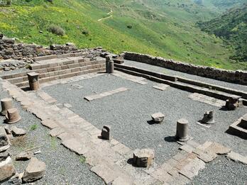 Ruins of an ancient stone structure sit on a gravel surface, surrounded by low stone walls and scattered columns, overlooking a lush, hilly landscape.