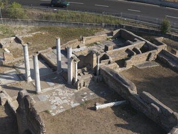 Aerial view of ancient stone ruins with several standing columns, broken walls, and scattered stone slabs, bordered by a fence with a road and car visible in the background.