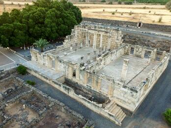 Aerial view of an ancient stone synagogue ruin surrounded by greenery and dry fields, with remnants of walls and columns visible, located near a tree-lined area and dirt pathways.