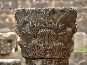 A weathered stone capital featuring carved vegetal motifs and a prominent menorah symbol on top, situated outdoors against a background of rough stone blocks.