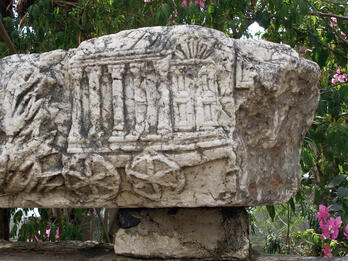 A weathered stone block with a carved relief depicting a chariot with columns and an arched door, set outdoors with foliage and flowers in the background.