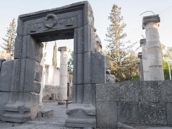 Ancient stone ruins with a large rectangular archway and several standing columns, surrounded by trees and greenery under a clear sky.