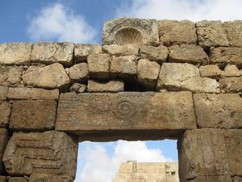 Stone doorway of an ancient ruin, featuring weathered blocks with decorative carvings, including a shell motif at the top and geometric patterns, against a partly cloudy sky.