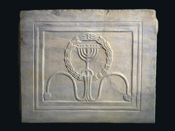 A carved marble tablet featuring a menorah inside a wreath, with two curved lines ending in leaf shapes beneath it. The design is framed by a rectangular border.