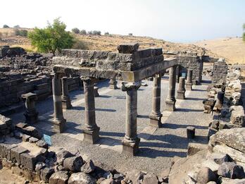 Ancient stone ruins of a rectangular structure with several columns and partial walls, set in a dry, hilly landscape with sparse vegetation under a clear sky.
