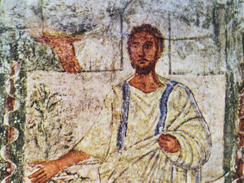 Ancient fresco of a bearded man in a white robe with blue stripes, standing beside a leafy bush, with his right arm extended and left hand holding his garment. The background shows architectural details and faded colors.