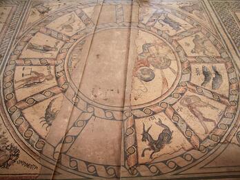 Ancient mosaic depicting the twelve zodiac signs arranged in a circle, each sign illustrated with a symbolic figure or animal, surrounded by detailed decorative borders. In the center of the circle is a person with a halo holding an orb.