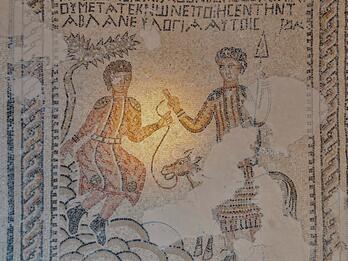 Ancient mosaic depicts two robed figures and a donkey. Greek text appears above the scene. There are areas of visible wear and missing tiles.