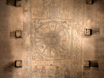 Mosaic panel with zodaic in the center, figures on the bottom, and ritual objects and animals on the top, surrounded by stone and several square blocks.