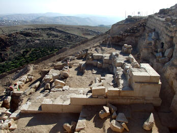 Ancient stone ruins at an archaeological site on a hillside, with scattered blocks and partial walls. Rolling hills and a distant settlement are visible in the background under a hazy sky.
