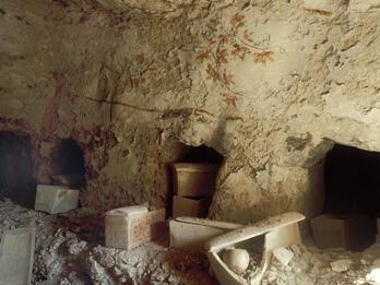 Ancient rock-cut tomb chamber with three burial niches in a rough limestone wall, several broken stone sarcophagi, and scattered debris on the ground. Faint painted decorations are visible on the wall.