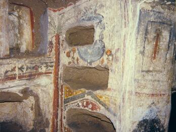 Ancient stone tomb with rectangular niches carved into the walls, decorated with faded frescoes and geometric patterns in red, yellow, and blue. Some niches appear empty; white mineral streaks run down the wall.