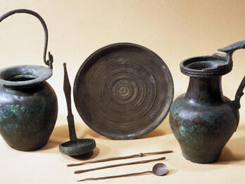 A collection of ancient bronze vessels and utensils, including two jugs with handles, a round plate, a ladle, and three long utensils.