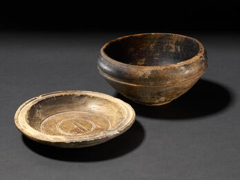 Two ancient wooden bowls on a dark background: one is deep and rounded with a blackish exterior, and the other is shallow and beige with a circular design etched inside.