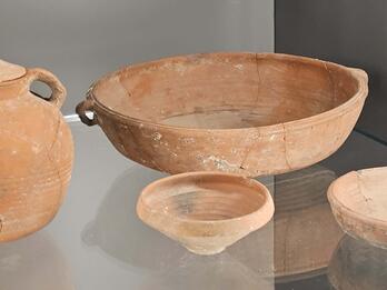 Four ancient, weathered ceramic vessels: a lidded jar with handles, a large bowl, a small bowl, and a shallow plate, displayed on a glass shelf against a plain, neutral background.