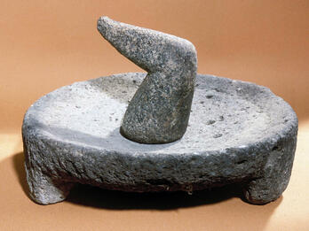 A grinder with a flat, round base and a hand-held cylindrical stone. The objects are gray and sit on a light brown surface.