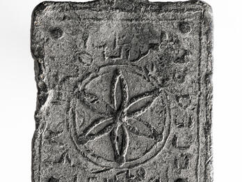 A rectangular, weathered metal object with a central flower-like geometric pattern inside a circle, surrounded by worn Hebrew inscriptions, with a hole at the top for hanging.