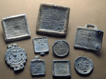 Nine ancient lead weights with various shapes, engravings, and inscriptions are arranged on a flat, brown surface. Some feature symbols, while others display text.
