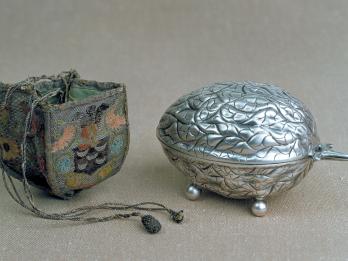 Silver oval container of decorative leaf design and woven, embroidered case next to it.