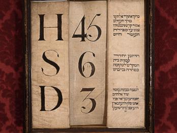 Framed piece of paper with three columns of text: single letters in left column, numbers in middle column, and Hebrew text in right column. 