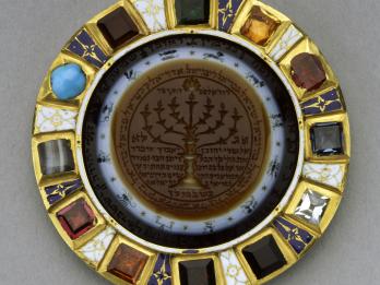 Circular amulet with stones around perimeter and image of candelabrum with Hebrew text in center, surrounded by images of the zodiac.