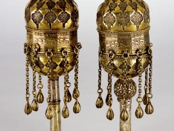 Two finials engraved with designs and Hebrew inscription, with chains hanging down from bottom and hand-shaped sculpture on top. 