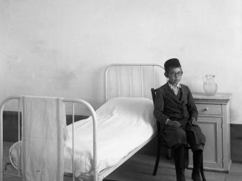 Photography of young boy sitting next to bed and nightstand.