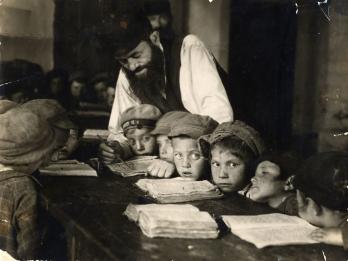 Photograph of young boys sitting at a long table with books open as teacher leans over them to teach.