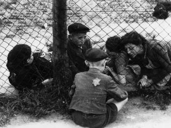 Photograph of women and children in ghetto facing photograph on the far side of a fence, interacting with another child on opposite side of fence with Star of David badge on his back.