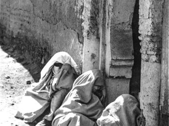 Photograph of three women seated on the floor next to building wearing hooded robes and veils and angled away from the viewer.