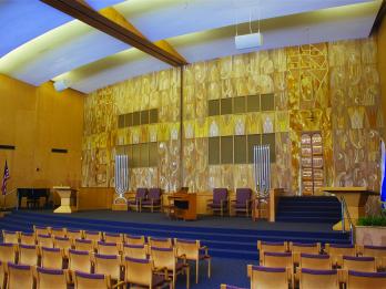 Photograph of interior view of a synagogue with rows of chairs facing a central platform featuring wall mural of painted panels with crowns, suns, Hebrew lettering, and spirals.