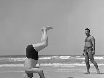 Photograph of two shirtless men on the beach, one doing a headstand with legs bent facing to the right of the viewer and the other man facing the viewer on the right side of the photograph.