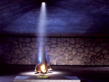 Memorial depicting flame burning from a base and illuminating a wall with an angular roof and basalt walls, with place names in English and Hebrew on the floor.