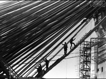 Photograph of men silhouetted on a steep ladder that bisects the photograph on a diagonal, below cables that run across the photograph, and tall building in background.