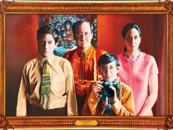 Photorealist painting of woman, man, and two young boys, one of whom holds camera, standing in front of a picture of a meditating god and looking at the viewer. 