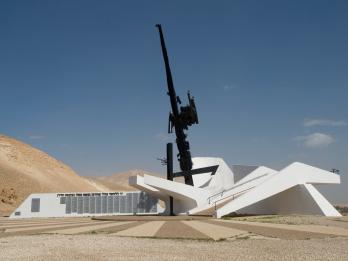 Cement and steel monument resembling a large firearm in a white base next to a building, surrounded by sand dunes.