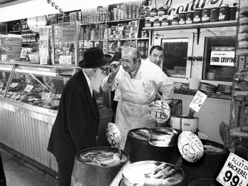 Photograph of interior of store with fish behind a glass counter and foodstuffs on shelves, with a man in a butcher's apron in center talking to a bearded man in a suit next to several barrels of fish and another man behind them.