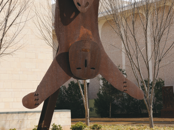 Steel sculpture on steel frame outside of building. Top of sculpture is the head of a sheep with horns and bottom of sculpture is stylized head and arms of a human figure.
