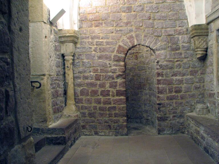 Photo of a stone room with an arched doorway, stone benches and columns, and a stairway.