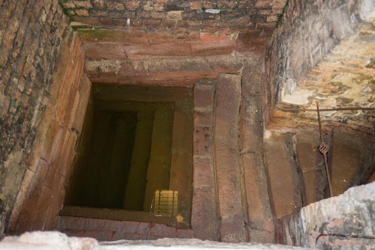 Photo of a rectangular stone pool, partially filled with water, with steps leading down into it, weathered brick and stone walls, and and stairs leading up through a doorway.