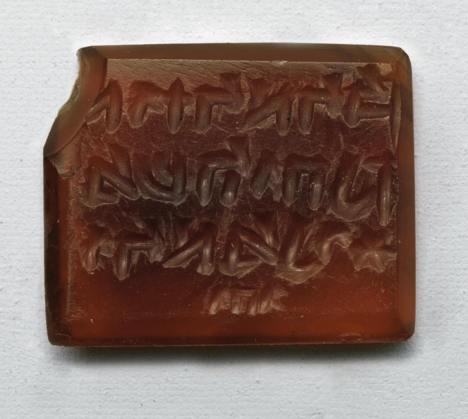 Photo of a rectangular red stone block with a beveled edge and chipped corner, inscribed with reversed Hebrew writing in three lines followed by a small word.