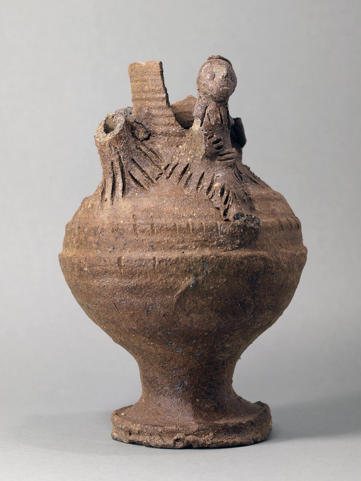 Earthenware vessel with a round body and flared base, with sculpted elements, some broken off, at the top, including a humanoid figure.