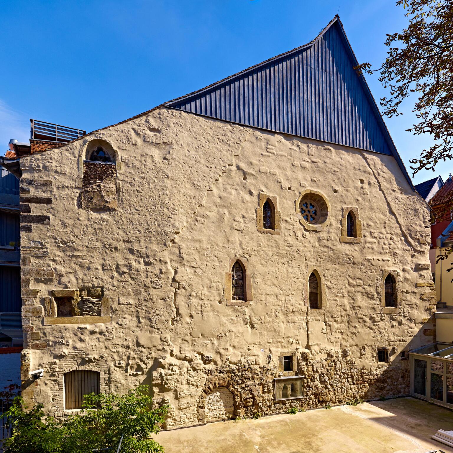 Photo of a beige stone building with Gothic arched windows, a circular window, a blue peaked roof, and irregular and patched stonework and other openings.