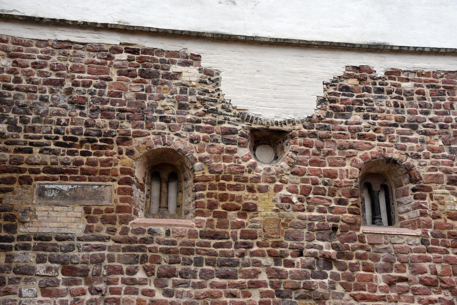 Photo of a weathered stone wall with two arched windows, a small circular window, several irrecular patches, and white-plastered brick along the top.